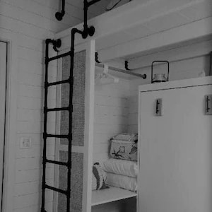 May include: A black metal ladder leading to a loft bed with a white wooden frame. The loft bed has a white closet with a sliding door and a white cabinet with a door. The closet has a shelf with a white blanket and a white storage bin.