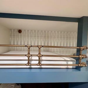 May include: A bunk bed with a brass pipe railing. The bed frame is painted blue, and the mattress is white. The headboard has a patterned wallpaper design. A brass fixture is mounted on the wall.
