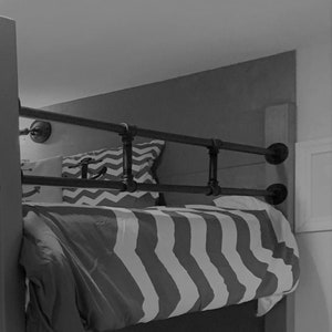 May include: Black metal pipe railing with black flanges attached to a wall, above a striped bed with a white and grey patterned pillow.
