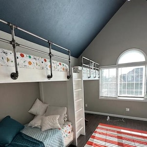 May include: A white bunk bed with a metal railing and a blue and white patterned duvet on the top bunk. The room has a grey and blue painted ceiling and a large window with white blinds.