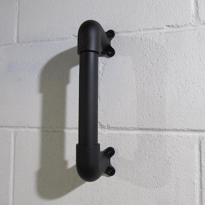 May include: A black metal pipe fitting, mounted vertically on a white breeze block wall. The fitting has a curved top and bottom with a straight section in the middle. Each end has a mounting bracket.