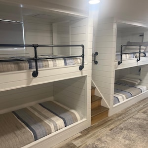 May include: A contemporary bunk bed arrangement featuring white shiplap walls and black pipe railings. The beds are dressed with striped bedding in blue, grey, and beige tones. Wooden steps provide access to the upper bunks, and the flooring is a light wood-effect laminate.