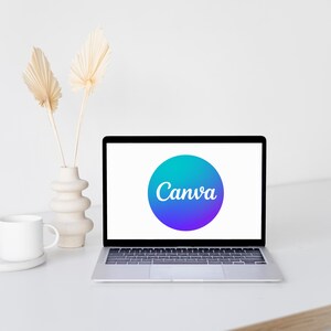 May include: A laptop displaying the Canva logo on its screen, set on a white desk. A white mug and a decorative vase with dried plants are to the left of the laptop. The Canva logo is a blue and purple gradient circle with white text.