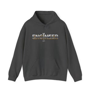 Puede incluir: Sudadera con capucha gris oscuro con bolsillo delantero y capucha con cordón. La palabra "ENGINEER" está impresa en el pecho en una fuente metálica en 3D, con un gráfico de llave inglesa incorporado.