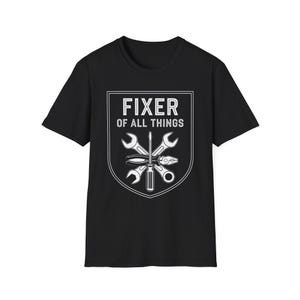 Puede incluir: Camiseta negra con un gráfico blanco de herramientas dentro de un escudo. El texto "FIXER OF ALL THINGS" está encima de las herramientas. El gráfico incluye llaves, un destornillador y otras herramientas de reparación.