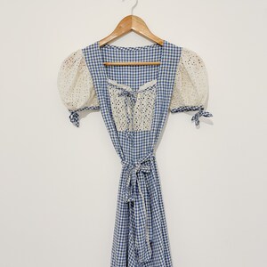 Puede incluir: Un vestido de cuadros azules y blancos con un corpi&ntilde;o de encaje blanco y mangas abullonadas. El vestido tiene un lazo en la cintura y un escote cuadrado.