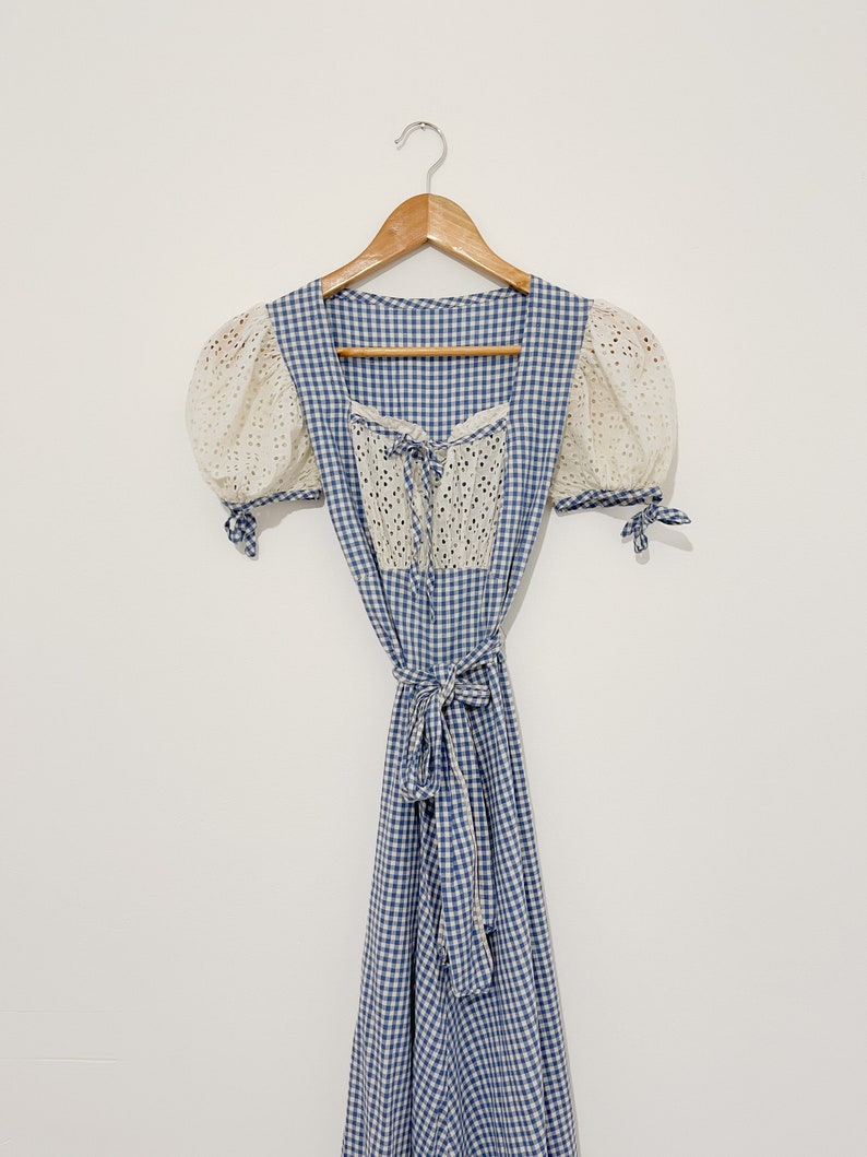 Puede incluir: Un vestido de cuadros azules y blancos con un corpi&ntilde;o de encaje blanco y mangas abullonadas. El vestido tiene un lazo en la cintura y un escote cuadrado.