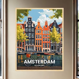 May include: A framed wall art print depicting a canal scene in Amsterdam, Netherlands. The illustration features colourful narrow houses, green trees, a bicycle in the foreground, and a canal with boats. The text AMSTERDAM NETHERLANDS is printed.