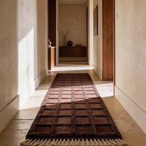 May include: A long, rectangular brown rug with a grid pattern and fringed edges. The rug is placed in a hallway with light-colored walls and flooring. The rug's texture and color add warmth to the space.