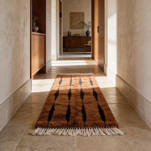 Könnte beinhalten: Ein rechteckiger, brauner Teppich mit einem schwarzen Rautenmuster und weißen Fransen. Der Teppich liegt in einem Flur mit Steinboden und Holztüren und -schränken. Der Teppich ist etwa 152 cm lang.
