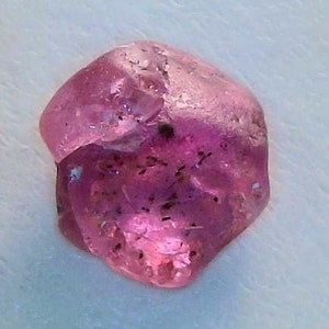 May include: A small, faceted, pink gemstone with dark inclusions. The gem is next to a quarter coin for size comparison, set against a white background. The gemstone has a rough, natural appearance.