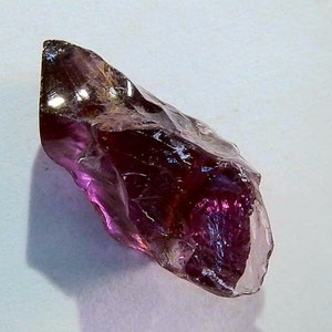 May include: A raw, translucent gemstone with a deep purple and magenta hue. The stone features an irregular, jagged shape with multiple faceted surfaces that catch the light, creating a glossy, reflective appearance across its crystalline structure.