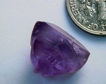 May include: A faceted, translucent amethyst gemstone with a deep purple hue. The gemstone is next to a US dime coin for size comparison. The amethyst has a crystalline structure and a polished surface, reflecting light.
