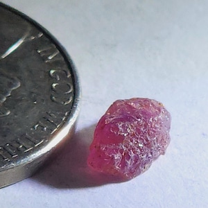 Puede incluir: Una gema de rubí en bruto, sin cortar, de color rosa intenso. El rubí está junto a una moneda para comparar el tamaño. La gema tiene una superficie texturizada y parece natural. La moneda muestra las palabras "IN GOD WE TRUST".