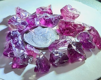 May include: A collection of irregularly shaped, translucent, deep pink crystals scattered around a quarter coin. The crystals have a faceted appearance, catching the light and displaying varying shades of pink and purple.