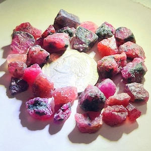 May include: A collection of raw, unpolished red ruby gemstones scattered around a silver-coloured coin. The stones exhibit varying shades of deep pink and crimson with irregular, jagged surfaces and dark mineral inclusions throughout the cluster.