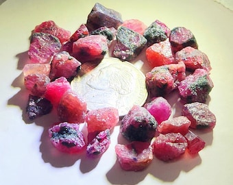 Könnte beinhalten: Eine Ansammlung von rohen, ungeschliffenen roten Rubinedelsteinen, die um eine silberfarbene Münze herum verstreut sind. Die Steine weisen verschiedene Schattierungen von tiefem Rosa und Karmesinrot mit unregelmäßigen, gezackten Oberflächen auf.