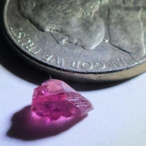 May include: A small, faceted, raw ruby gemstone with a deep pink hue. The gemstone is next to a silver coin, providing a size reference. The coin has the words "IN GOD WE TRUST" and "LIBERTY" visible.