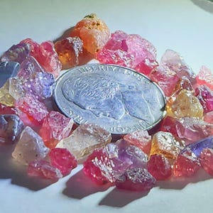 May include: A collection of raw gemstones in shades of pink, orange, yellow, and purple surrounds a silver quarter coin. The gemstones are rough-cut and have a natural, unpolished appearance, with varying sizes and shapes.