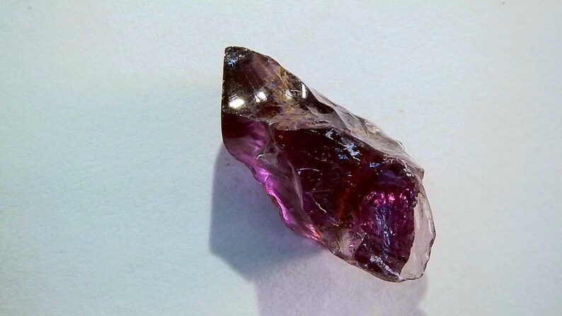May include: A raw, translucent gemstone with a deep purple and magenta hue. The stone features an irregular, jagged shape with multiple faceted surfaces that catch the light, creating a glossy, reflective appearance across its crystalline structure.
