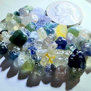 Peut inclure: Un ensemble de pierres précieuses brutes de différentes couleurs, notamment bleu, vert, jaune et transparent, disposées sur une surface blanche. Une pièce de monnaie argentée est présente pour l'échelle. Les pierres ont une texture naturelle et rugueuse.