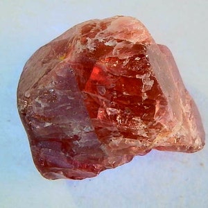 May include: A rough, uncut gemstone with a reddish-orange hue. The mineral has a crystalline structure and a slightly translucent appearance. The stone is set against a white background.
