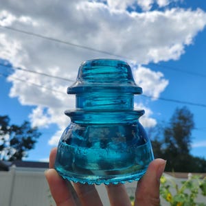 May include: A vintage, turquoise-colored glass insulator. The insulator has a tiered design with a rounded top and a flared base. The glass is transparent, allowing light to pass through, and is held against a backdrop of a bright blue sky with fluffy white clouds.