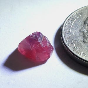 Puede incluir: Una pequeña gema de color rojo rubí, de corte tosco, con una superficie facetada. La gema está junto a una moneda para comparar el tamaño. La gema tiene un aspecto natural, sin pulir, con una calidad ligeramente translúcida.