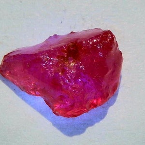 May include: A raw, translucent gemstone in a rich red colour. The stone's surface is slightly irregular, reflecting light and creating highlights. This gem is likely a ruby, often used in jewellery.