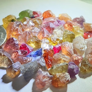 Może przedstawiać: Kolekcja surowych, nieoszlifowanych kamieni szlachetnych w różnych kolorach, w tym czerwonym, pomarańczowym, żółtym i fioletowym. Moneta służy jako odniesienie do rozmiaru. Kamienie szlachetne mają nieregularne kształty i są przezroczyste.