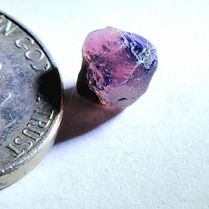 May include: A close-up of a small, rough-cut gemstone with a mix of pink and blue hues. The gemstone is next to a silver coin, providing a size reference. The stone has a natural, unpolished appearance.