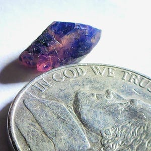 May include: A small, raw gemstone with a mix of purple and pink hues. The gem is positioned next to a silver coin, providing a size reference. The coin's inscription reads "IN GOD WE TRUST".
