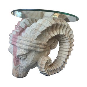 May include: A sculpted ram's head with a glass top table. The ram's head is made of a light beige stone with pink accents. The glass top is clear and round.