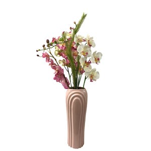 May include: A blush pink vase with a fluted design holds a vibrant arrangement of artificial flowers. The arrangement includes white and pink orchids, and green accents. The vase is a tall, elegant shape.