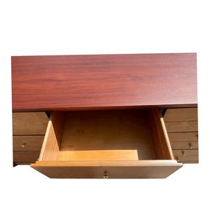 May include: A wooden desk with a dark reddish-brown top and a drawer pulled open. The drawer is made of light-colored wood and has a brass knob. Several other drawers are visible on the sides of the desk.
