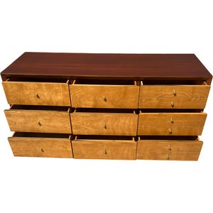 May include: A mid-century modern dresser with nine drawers. The top is a dark brown, and the drawers are light wood with brass knobs. All drawers are open, revealing the interior.