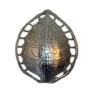 May include: A silver turtle shell shaped serving dish with a detailed design. The dish is made of metal and has a cutout design around the edge.