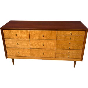 May include: A mid-century modern dresser with a dark brown top and a light wood body. The dresser has nine drawers with gold-colored knobs and tapered legs. The wood grain is visible on the drawers.