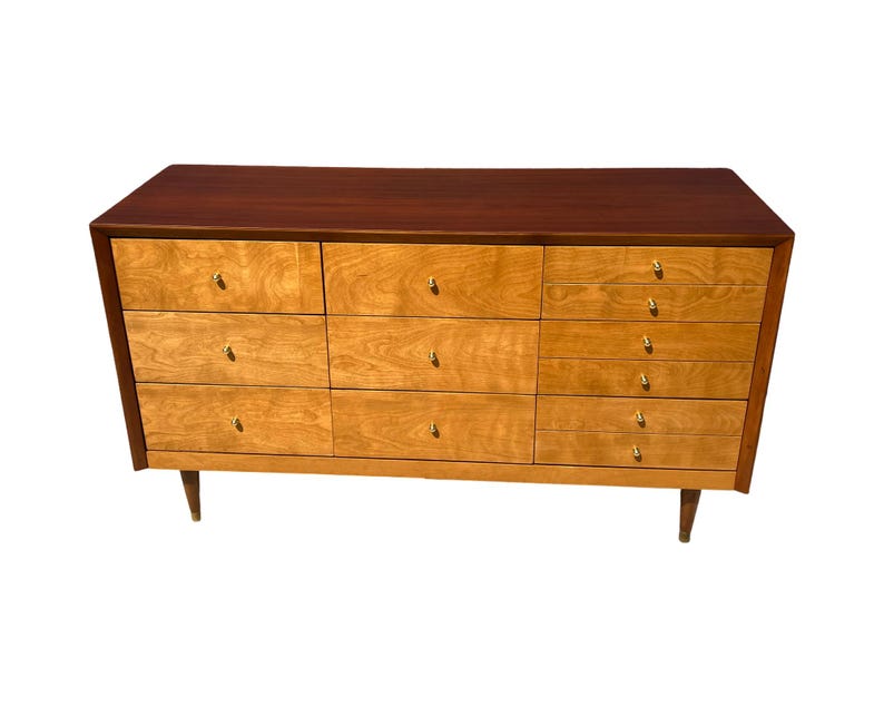 May include: A mid-century modern dresser with a dark brown top and a light wood body. The dresser has nine drawers with gold-colored knobs and tapered legs. The wood grain is visible on the drawers.