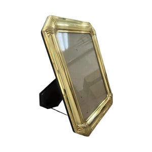 May include: A gold-colored picture frame with an ornate, detailed border. The frame is rectangular and has a black easel back for display. The frame is angled slightly, showcasing its design and the space for a photo.