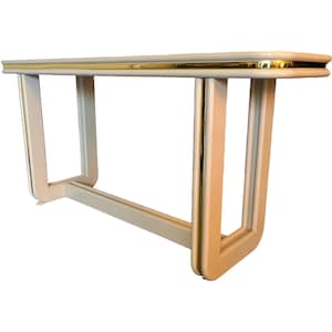 May include: A cream-colored console table with a gold trim. The table has a rectangular top and a unique open-frame design with curved legs. The gold trim accents the edges of the table, adding a touch of elegance to the furniture piece.