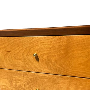 May include: Close-up of a wooden dresser with a light brown finish. The drawers have a natural wood grain pattern and are accented with small, gold-colored knobs. The top edge is a darker brown.