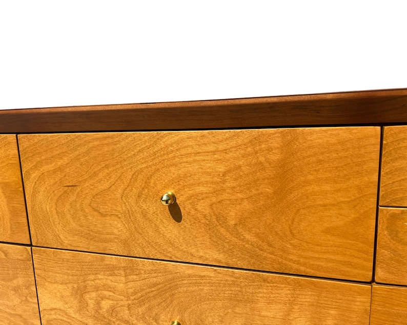 May include: Close-up of a wooden dresser with a light brown finish. The drawers have a natural wood grain pattern and are accented with small, gold-colored knobs. The top edge is a darker brown.