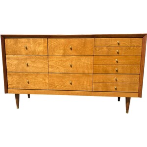 May include: A mid-century modern dresser with a light wood finish and a dark wood trim. The dresser has multiple drawers with gold-colored knobs and tapered legs. The design is simple and elegant, suitable for a bedroom or living space.
