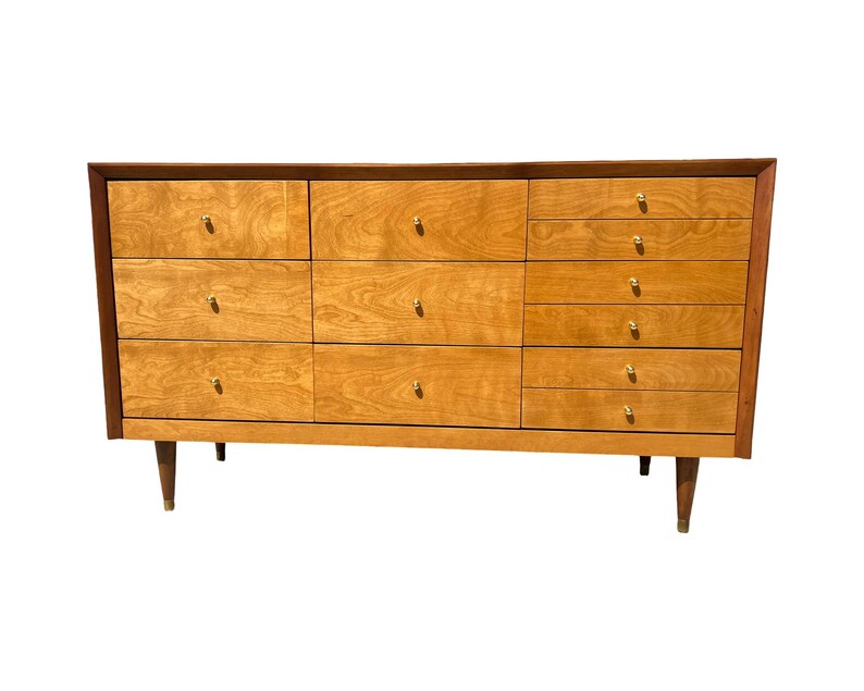 May include: A mid-century modern dresser with a light wood finish and a dark wood trim. The dresser has multiple drawers with gold-colored knobs and tapered legs. The design is simple and elegant, suitable for a bedroom or living space.