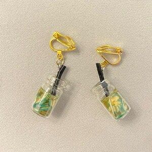 May include: A pair of clip-on earrings featuring miniature clear glass beverage cups with yellow and green accents, and black straws. The earrings have gold-colored clip-on closures.