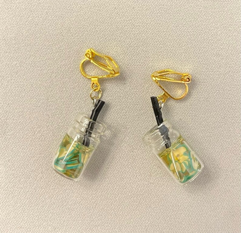 May include: A pair of clip-on earrings featuring miniature clear glass beverage cups with yellow and green accents, and black straws. The earrings have gold-colored clip-on closures.