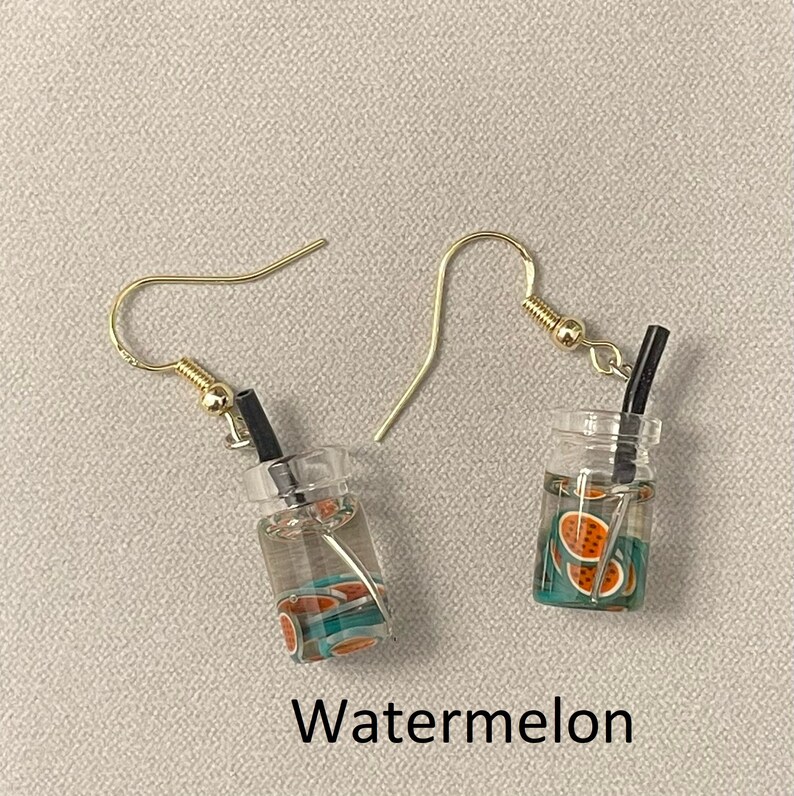 May include: A pair of gold-colored hook earrings. Each earring features a miniature clear glass jar with a black straw and watermelon slices. The word "Watermelon" is printed in black at the bottom.