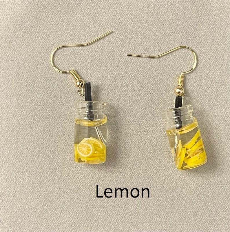 May include: A pair of novelty earrings featuring miniature clear bottles filled with yellow lemon slices and a black straw. The earrings have gold-colored fish hook ear wires. The word "Lemon" is printed in black text below the earrings.