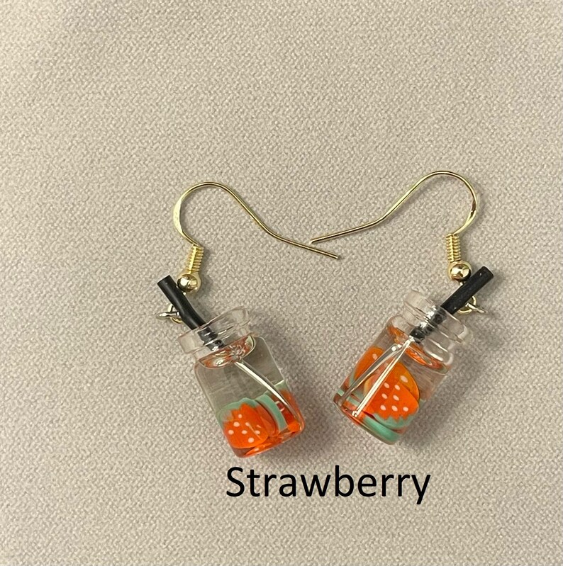 May include: A pair of dangle earrings featuring miniature clear glass bottles with a strawberry design. Each earring has a gold-colored hook and a black straw. The word "Strawberry" is printed below the earrings.